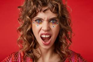 Excited blue-eyed curly young female with multicolored dots on her face looking at camera with wide mouth opened and frowning her eyebrows, isolated over red background photo