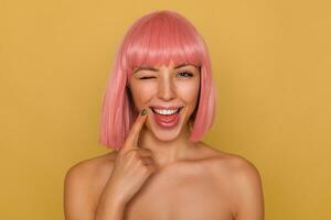 Joyful young attractive pink haired female with bob hairstyle holding index finger on her face while smiling broadly and giving happily wink to camera, posing over mustard background photo