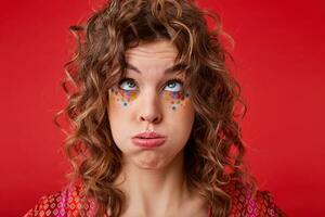 Funny portrait of pretty young female with curly hair and festive makeup looking upwards and blowing on her hair, making fun while posing over red background, wearing motley patterned top photo