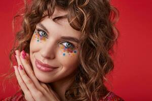 Positive pretty young female with romantic hairstyle and multicolored dots on her face looking at camera with tender smile and leaning chin on folded hands, standing over red background photo