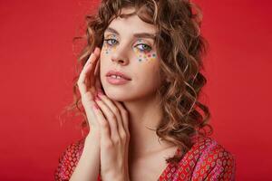 Delicate pretty lady with romantic hairstyle and multicolored dots on her face wearing motley patterned top while posing over red background, folding hands near face and looking gently to camera photo