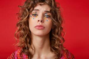 Upset young curly female with festive makeup wearing motley patterned top, frowning and looking to camera with folded lips, posing over red background photo