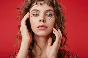 Beautiful blue-eyed curly woman with multicolored dots on her face wearing motley patterned top, keeping hands gently near her face, isolated over red background photo