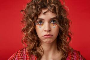 estudio foto de triste de ojos azules joven mujer con romántico peinado y festivo maquillaje mirando a cámara lamentándose, posando terminado rojo antecedentes en abigarrado estampado parte superior