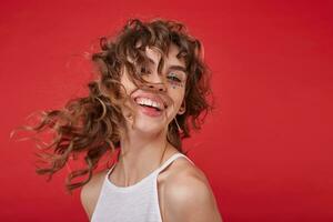 retrato de contento joven bonito mujer con Rizado pelo y festivo hacer p riendo alegremente y mirando a cámara, posando terminado rojo antecedentes en blanco camisa foto