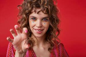 Coquettish young curly woman with multicolored dots on her face looking at camera and biting underlip, raising hand playfully while standing over red background in motley patterned top looking camera photo