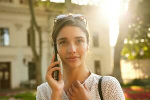 de cerca de atractivo joven oscuro peludo mujer con Gafas de sol en su cabeza mirando en frente de su mientras hablando en teléfono, caminando terminado ciudad antecedentes en soleado calentar día foto