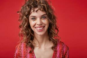 Beautiful cheerful young lady with curly hair and festive makeup standing over red background in motley patterned top, looking at camera with happy wide smile photo