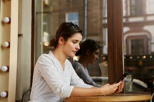 Side view of young pretty brunette woman with bun hairstyle sitting over cafe interior with mobile phone in her hands and looking on screen, dressed in white polka-dot dress photo