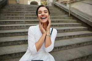 Overjoyed young attractive brunette lady with casual hairstyle holding face with raised hands and looking happily to camera, sitting over city stairs in white polka-dot dress photo