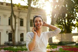 agradable mirando joven morena mujer con Gafas de sol en su cabeza teniendo bonito conversacion en teléfono y sonriente felizmente a cámara, caminando a lo largo calle en soleado día foto