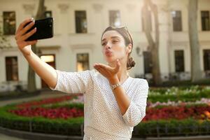 Beautiful young dark haired woman with casual hairstyle posing outdoor on warm spring day with mobile phone in raised hand, blowing air kiss to camera while making photo of herself