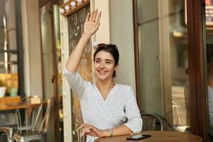 Positive young dark haired female looking aside and smiling happily while raising hand in hello gesture, meeting friends in city cafe on sunny weekend day photo