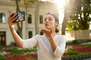 al aire libre foto de atractivo joven morena hembra con bollo peinado enviando aire Beso a cámara mientras haciendo selfie con teléfono inteligente, en pie terminado urbano ambiente en calentar brillante día