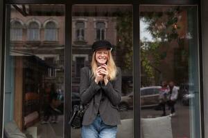 alegre joven encantador rubia largo peludo dama participación taza de café en elevado manos y riendo felizmente con cerrado ojos, posando al aire libre terminado grande ventana en de moda ropa foto