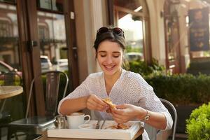 Pleasant looking young dark haired woman with casual hairstyle having tasty breakfast outdoor on sunny morning, being in nice mood and smiling cheerfully photo