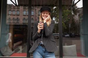 al aire libre Disparo de agradable mirando contento joven rubia dama sonriente alegremente con cerrado ojos y participación palma en su mejilla, en pie terminado café exterior con papel taza foto