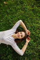 Attractive young dark haired brunette woman in white polka-dot clothes lying on green grass with hands under her head, looking positively at camera with broad smile photo