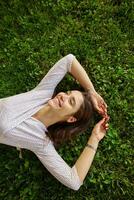 Pleased young long haired brunette female in elegant clothes relaxing on green lawn on warm spring day, keeping hands raised and smiling pleasantly while looking aside photo