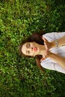 Portrait of young pretty long haired brunette female raising palms and folding lips in air kiss while looking positively at camera, posing over green grass in elegant clothes photo