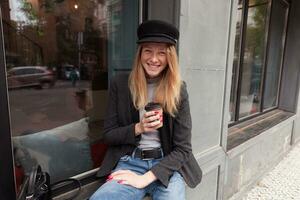 alegre atractivo joven rubia largo peludo mujer en de moda ropa sentado terminado grande café ventana con taza de café en su mano, mirando a cámara con amplio contento sonrisa foto