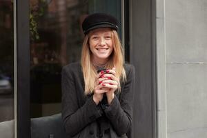 retrato de hermosa positivo joven rubia mujer en negro sombrero participación taza de café en elevado manos y mirando alegremente a cámara con encantador sonrisa, posando al aire libre en gris elegante ropa foto