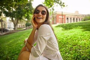 Cheerful attractive young dark haired female in sunglasses holding hand on her face while laughing joyfully, sitting over city background on warm sunny day photo