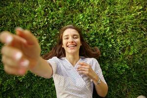 Cheerful young long haired brunette lady in white polka-dot dress laughing happily while looking at camera, raising hand to camera while lying on green grass photo