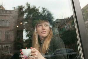 Disparo de joven bonito rubia largo peludo mujer con natural maquillaje participación blanco cerámico taza en su manos mientras mirando pensativamente a ventana, vestido en calentar elegante ropa foto