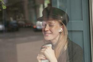 agradable mirando joven de moda dama con rubia largo pelo sentado terminado antepecho en ciudad café y acuerdo teléfono inteligente en su manos, sonriente atentamente con cerrado ojos foto