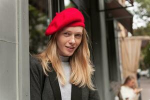 de cerca de hermosa joven mujer con largo rubia pelo vistiendo elegante ropa mientras posando terminado urbano fondo, mirando a cámara y sonriente ligeramente foto