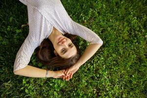 Portrait of lovely young long haired brunette lady keeping hands raised while looking positively at camera with charming smile, posing over green grass on warm bright day photo