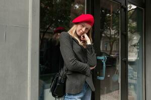 Oudoor shot of bored young blonde long haired woman standing over cafe exterior and leaning her head on raised hand, dressed in grey coat, jeans and red beret photo