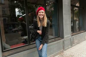 alegre joven atractivo rubia dama vistiendo de moda ropa mientras caminando terminado ciudad y siendo en alto espíritu, mirando felizmente a cámara con amplio sonrisa foto