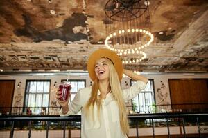 Indoor shot of happy pretty young blonde female with long hair dressed in trendy clothes posing over city cafe interior, holding her hat and looking aside joyfully, laughing about funny joke photo