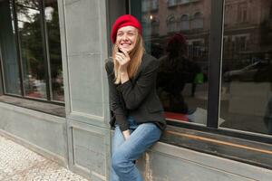 alegre joven encantador rubia largo peludo mujer sentado al aire libre terminado antepecho y levantamiento mano a su cara mientras riendo felizmente, vestido en elegante ropa foto