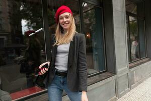 Positive young attractive lady wearing her blonde hair in casual hairstyle while walking over street with her friends, being in nice mood and smiling cheerfully photo