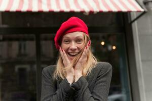 Close-up of overjoyed young blonde lady with red manicure holding her face with raised hands and looking surprisedly to camera, laughing happily while standing over cafe exterior photo