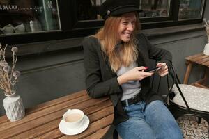 al aire libre Disparo de contento hermosa joven rubia mujer vestido en de moda ropa teniendo desayuno en café terminado verano terraza, sonriente felizmente mientras mecanografía mensaje en su teléfono foto