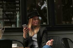 Pensive young attractive blonde female with casual hairstyle posing over cafe exterior in elegant clothes, looking aside with folded lips and holding mobile phone photo