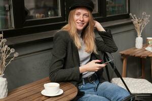 al aire libre foto de joven de ojos azules rubia hembra con rojo manicura acuerdo móvil teléfono en su mano y mirando afirmativamente a cámara mientras posando terminado ciudad café en soleado día