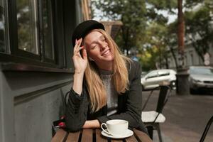 satisfecho joven encantador rubia hembra con natural maquillaje sentado a mesa terminado ciudad café y Bebiendo café, disfrutando soleado día con cerrado ojos y sonriente atentamente foto