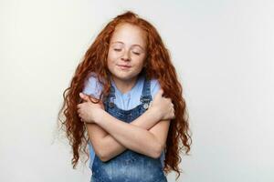 Portrait of cute freckles little girl with ginger hair, hugs himself and dreaming about puppy with closed eyes, stands over white background and dreamily smiling. photo