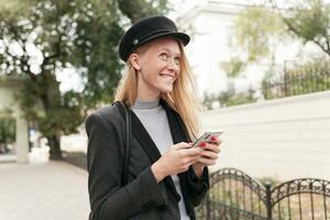 soñador joven positivo rubia mujer con rojo manicura mordiendo debajo del labio mientras mirando alegremente hacia arriba y sonriente atentamente, acuerdo móvil teléfono en elevado manos mientras posando al aire libre foto