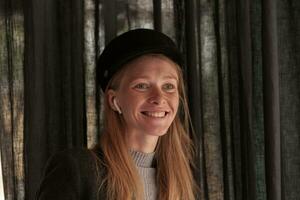 Portrait of beautiful young blonde long haired female in black hat wearing headphones while posing over cafe interior, being in nice mood and smiling positively photo