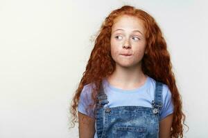 Portrait of curious cute freckles little girl with ginger hair, thinking about something, bites lips, looks away over white background with copy space on the left side. photo