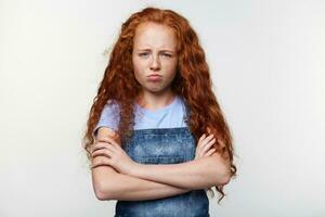 Prtrait of unhappy freckles little girl with ginger hair, stands over white wall with crossed arms, looks sad and offended. photo