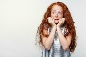 Scared little girl with ginger hair and freckles, afraid and anxious biting her finger nails, looking at camera with wide opened eyes and looks away, isolated over white background with copy space. photo