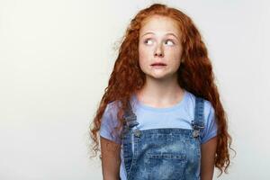 Portrait of guilty cute freckles little girl with ginger hair, pretends not to do anything wrong, looks away over white background with copy space on the left side. photo