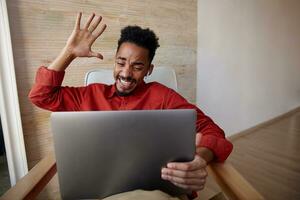 Indoor photo of excited young pretty short haired brunette guy with dark skin keeping his hand raised while looking emotionally on his laptop, sitting over beige interior